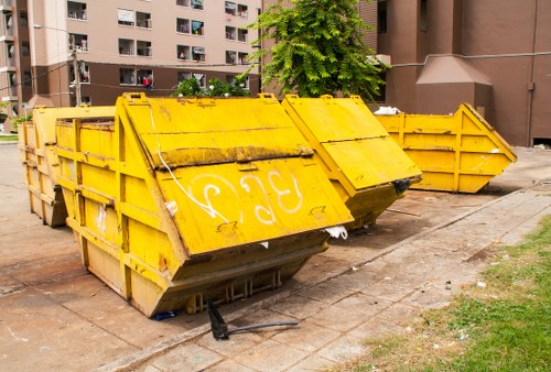 A row of large, heavy-duty yellow metal skips positioned on a paved urban street, with one skip slightly tilted forward and the others set in a line behind it. The skips have reinforced sides and are used for waste collection, showing signs of dirt and weathering with some rust around the edges. The front skip's lid is closed, partially obscuring the interior, which appears to contain mixed debris. In the background, there are multi-storey residential buildings with balconies and windows, along with a tree providing some greenery. The setting is during daylight hours, with natural light illuminating the scene. The area around the skips is clean but shows typical urban wear, including slight dirt on the pavement. This image relates to commercial waste removal services, such as those provided by Commercial Waste Removal Morden, and captures the typical outdoor waste container setup on the streets of Morden, Surrey, within the London Borough of Merton.