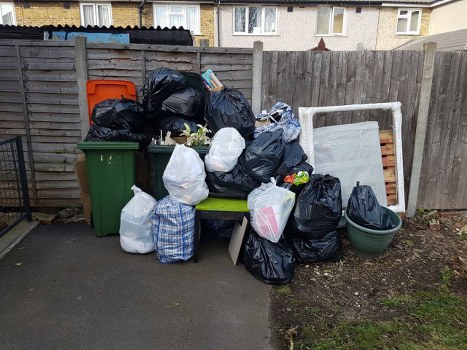 A collection of various black and white rubbish bags, including some with checked patterns, stacked and scattered on a paved outdoor area in front of a wooden garden fence. Among the bags, there is a large green wheelie bin, a smaller green planter, and a collapsed wooden bed frame with a white mattress leaning against the fence. The environmental setting appears to be a residential backyard or garden space in Morden, with a mix of cleared and unpaved ground visible. The scene suggests a rubbish removal situation, likely handled by Commercial Waste Removal Morden, with an emphasis on domestic waste disposal. Natural daylight highlights the different textures and colours of the waste bags and outdoor surroundings, creating a typical scene of refuse awaiting collection or disposal in a suburban setting.