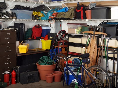 The image depicts a cluttered garage or storage area filled with various items typically associated with rubbish and household storage. In the foreground, there are multiple plastic plant pots of different sizes and colours, with some stacked and others placed individually on the concrete floor. A blue children's bicycle with black wheels and orange accents is leaning against a shelving unit on the right side. The shelving comprises several levels, holding an assortment of containers, boxes, and miscellaneous objects such as plastic storage bins, a black toolbox, and a red fire extinguisher. Mounted above on the wall are white metal wire shelves loaded with various items including a yellow and red toolbox, rolled-up cables, gardening tools, and a black case. The environment appears to be a typical home garage used for storing tools, cleaning supplies, and miscellaneous clutter, with a neutral lighting that suggests indoor illumination. The scene is indicative of a space that could benefit from professional rubbish removal services like those offered by Commercial Waste Removal Morden, serving the Morden area with comprehensive rubbish clearance solutions.