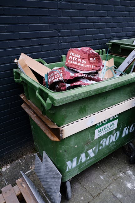 A black wheeled rubbish bin labeled with the name 'St. John's' is positioned on a sidewalk beside a curb in an outdoor urban environment at night. The bin is filled with various waste materials includ