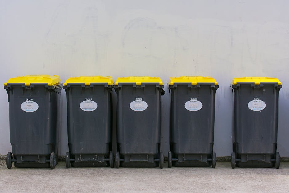 A row of five large wheeled rubbish bins with black plastic bodies and bright yellow lids, positioned against a plain, light grey concrete wall. The bins are aligned closely on a paved surface, with t