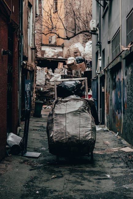 A street scene in an urban area shows a white waste collection truck with an open rear compactor, positioned close to the curb and partially extending into the roadway. The truck's surface appears sli