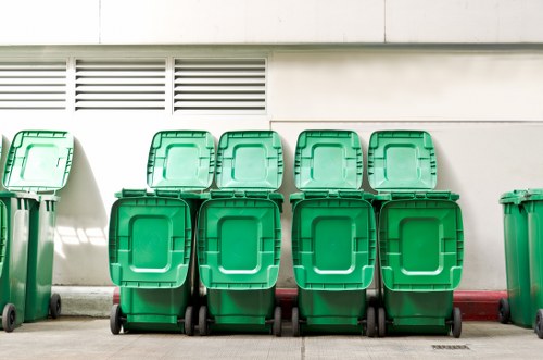 A row of eight large green wheeled rubbish bins with closed lids, positioned on a concrete driveway in front of a white building wall with horizontal ventilation slats. The bins are made of durable plastic with textured surfaces and slightly rounded edges, aligned in two rows of four. To the left, part of an additional green bin is partially visible. The scene appears to be outdoors, in a residential or commercial area, with the bins' wheels resting directly on the pavement. The positioning suggests they are ready for collection or disposal, consistent with waste management services in Morden or similar areas. The background includes the building's clean, smooth wall, and the overall setting is organised and tidy, reflecting a professional approach to rubbish collection by companies such as Commercial Waste Removal Morden.
